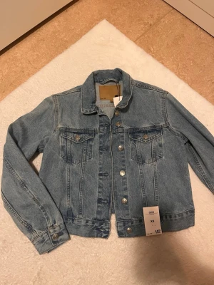 Ljusblå klassisk jeansjacka XS - Snygg ljusblå jeansjacka med klassisk passform och silvriga knappar. Jackan har två bröstfickor med lock och knappar samt raka sömmar för en tidlös look. Perfekt att slänga över en hoodie eller t-shirt för en cool stil.
