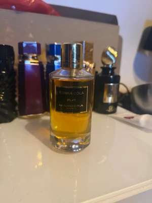 Mancera Tonka Cola 120ml EDP  - Mancera Tonka Cola Eau de Parfum i en elegant, cylindrisk glasflaska med guldfärgat lock och etikett. Doften har en djup bärnstensfärgad vätska och flaskan utstrålar lyxig och modern känsla. Perfekt för dig som gillar unika och intensiva parfymer.