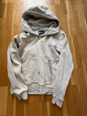 Beige hoodie med teddyfoder - Mysig beige hoodie med stor huva och teddyfoder på insidan. Har en halv dragkedja vid halsen och känguruficka framtill. Tillverkad i mjukt material och har ribbade muddar vid ärmslut och nederkant. Perfekt för kalla dagar.