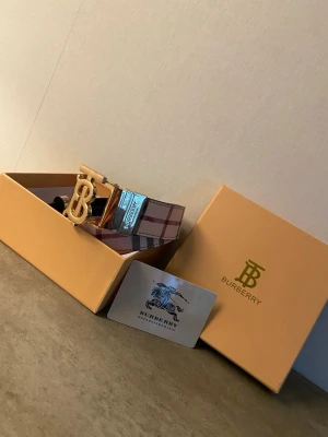 Rutig Burberry bälte med gulddetalj - Säljer ett klassiskt Burberry bälte med ikoniskt rutigt mönster i beige, rött och brunt. Bältet har ett stort guldfärgat BB-spänne som ger en lyxig känsla. Perfekt accessoar för att lyfta vilken outfit som helst.