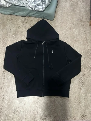 Svart hoodie från Polo Ralph Lauren - Säljer en svart hoodie med dragkedja från Polo Ralph Lauren. Klassisk design med huva och dragsnören, samt den ikoniska vita logon broderad på bröstet. Tillverkad i mjukt bomullsmaterial och har ribbade muddar vid ärmar och midja.