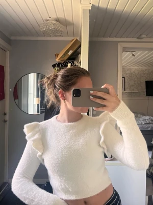 Vit fluffig stickad tröja med volanger - Supersöt vit stickad tröja med fluffig textur och volangdetaljer på axlarna. Tröjan har rund halsringning och långa ärmar. Perfekt för dig som vill ha något gulligt och trendigt i garderoben.