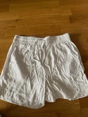 Vita linne liknande loose shorts med snörning - Säljer ett par vita loose shorts med elastisk midja och snörning framtill. Shortsen har sidofickor och en bakficka, tillverkade i ett tunt och luftigt tyg som passar perfekt till varma dagar.