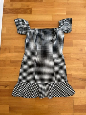 Rutig kortklänning från H&M Divided - Står storlek 40 men liten i storlek så passar 34/36 och xs/s🥰🥰Supersöt svartvit rutig kortklänning från H&M Divided med volangkant nertill och offshoulder-korta ärmar. Klänningen har en figurnära passform och är perfekt för sommarens alla tillfällen. Tillverkad i mjuk bomull för en skön känsla.