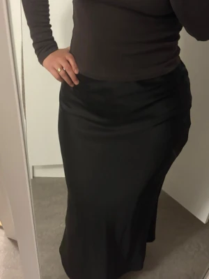 Svart långkjol i satin - Stilren svart långkjol i glansigt satinmaterial. Kjolen har en mjuk, följsam passform och faller snyggt längs benen. Perfekt att matcha med en enkel topp för en trendig look.