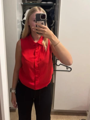 Röd ärmlös blus med krage - Snygg röd blus i ärmlös modell med klassisk krage och knappar framtill. Tillverkad i ett lätt och luftigt material som passar perfekt till både jeans och kjol. Enkel att styla för en trendig look.