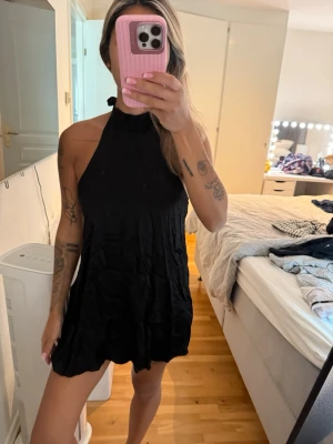 Svart halterneck kortklänning - Säljer en svart kortklänning med halterneck och lös passform. Klänningen är ärmlös och har en enkel, stilren design som passar perfekt till sommaren. Materialet ser ut att vara lätt och luftigt, vilket gör den skön att bära under varma dagar.