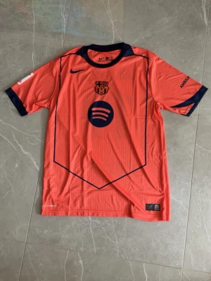 Barça Nike matchtröja orange - Säljer en orange FC Barcelona matchtröja från Nike med mörkblå detaljer och klubbmärke på bröstet. Tröjan har rund hals, korta ärmar och är tillverkad i Dri-FIT material som andas. Spotify-logga på framsidan och snygga grafiska linjer.
