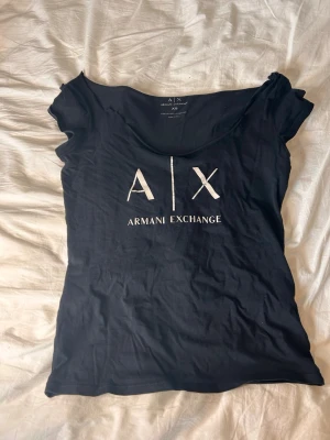 Svart t-shirt från Armani Exchange - Snygg svart t-shirt från Armani Exchange med stort vitt A|X-tryck på bröstet. T-shirten har rund halsringning och korta ärmar. Tillverkad i mjuk bomull som känns skön mot huden. Perfekt för en clean och stilren look.