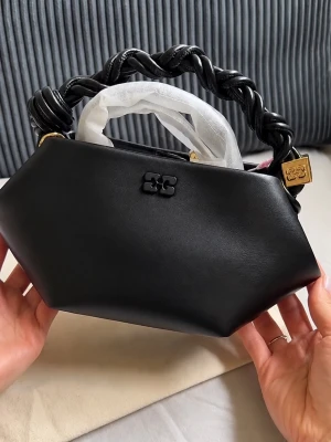 Svart geometrisk handväska med flätat handtag - Snygg svart handväska i geometrisk form med ett flätat handtag i läderimitation. Väskan har guldfärgade detaljer och en diskret logotyp framtill. Perfekt för dig som gillar unika accessoarer och vill sticka ut lite extra. Mått: 17 cm i höjd, 28 cm i bred, och 14 cm i djup. Kom med erbjudanden.