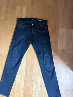 Svarta Replay Anbass jeans - Snygga svarta jeans från Replay, modell Anbass. Klassisk femficksdesign med smal passform och raka ben. Jeansen har knappgylf och är tillverkade i ett mjukt denimtyg som ger en skön känsla. Perfekta för dig som gillar stilrena och tidlösa jeans. Storlek 30,30