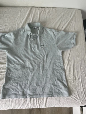Ljusgrå pikétröja från Lacoste - Klassisk ljusgrå pikétröja från Lacoste med den ikoniska broderade krokodilloggan på bröstet. Tröjan har korta ärmar, krage och knappar framtill. Perfekt för en clean och stilren look.