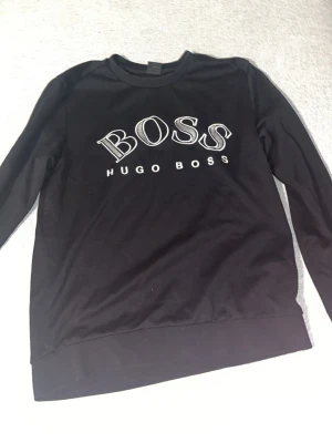 Svart stickad tröja från Hugo Boss - Svart stickad tröja från Hugo Boss med stor vit logga och texten 'BOSS HUGO BOSS' på bröstet. Tröjan har rund halsringning, långa ärmar och ribbade muddar vid ärmslut och nederkant. Enkel och stilren design som passar till mycket.