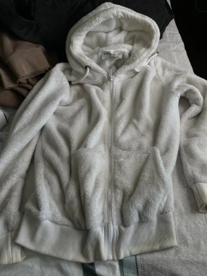 Vit fluffig hoodie från Amaci & Sons - Mysig vit hoodie från Amaci & Sons i storlek S. Tillverkad i supermjuk, fluffig fleece med dragkedja framtill, huva och breda ribbade muddar vid ärmslut och nederkant. Perfekt för chill dagar eller när du vill ha något riktigt bekvämt.