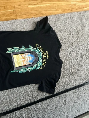 Svart t-shirt med Casablanca-tryck - Säljer en svart t-shirt med färgglatt tryck på både bröstet och ryggen. Motivet visar en port med gröna blad och texten 'Avenida Casablanca'. T-shirten har rund hals och korta ärmar. Perfekt för dig som gillar grafiska prints och streetwear.