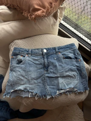 Blå jeanskjol med fransig kant från Nelly  - Säljer en blå jeanskjol i kort modell med rå, fransig kant nertill. Kjolen har klassiska fem fickor, bälteshällor och knappgylf. Perfekt för dig som gillar en avslappnad och trendig stil.