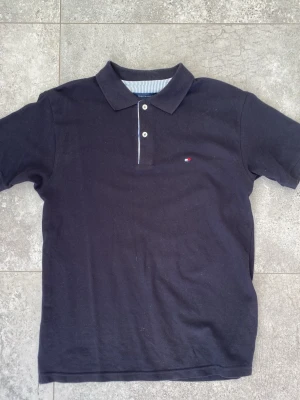 Svart piké från Tommy hilfiger - Klassisk svart pikétröja med krage och två knappar framtill. Tröjan har korta ärmar och en liten broderad detalj på bröstet. Perfekt till jeans eller shorts för en clean och enkel stil. Hör av er vid frågor✅