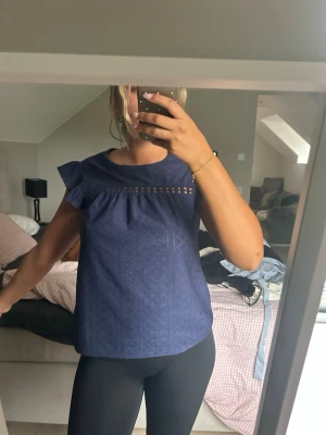 Blå blus med volangärmar - Söt blå blus med volangdetaljer på ärmarna och broderade hålmönster. Toppen har rund halsringning och en dekorativ öppen detalj över bröstet. Perfekt för dig som gillar en feminin och avslappnad stil.