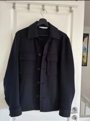Svart overshirt från ZARA - Stilren svart overshirt från ZARA med klassisk krage och stora bröstfickor med lock. Knäpps med fem knappar framtill och har raka ärmar med breda muddar. Perfekt lager-på-lager-plagg i ett mjukt och lite tjockare material.58%bomull ,2%elastan,40%polyster 