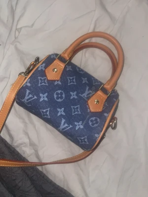 Blå monogram handväska i denim - Snygg liten handväska i blå denim med klassiskt monogrammönster och detaljer i ljusbrunt läder. Väskan har både handtag och en avtagbar axelrem i läder, samt guldfärgade metalldetaljer. Perfekt för dig som vill ha en trendig accessoar med lyxig känsla.