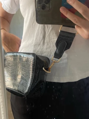 Svart croco-mönstrad handväska - Snygg svart handväska med croco-mönster och bred axelrem. Väskan har guldfärgade detaljer och dragkedja upptill. Materialet ser ut som konstläder med struktur som påminner om krokodilskinn. Perfekt för dig som vill ha en trendig accessoar.