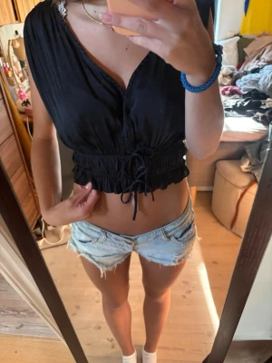 Svart croppad omlottopp med snörning - Säljer en svart croppad omlottopp med snörning i midjan. Toppen är ärmlös och har en djup v-ringning som ger en snygg och avslappnad look. 