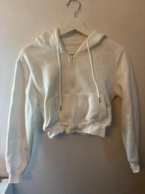 Vit croppad hoodie med dragkedja - Säljer en vit croppad hoodie med dragkedja framtill och två fickor. Hoodien har huva och ribbade muddar vid ärmar och midja. Materialet känns mjukt och bekvämt.