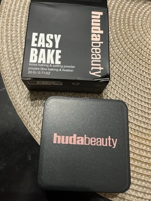 Huda Beauty Easy Bake Peach Pie - Loose baking & setting powder från Huda Beauty i nyansen Peach Pie. Kommer i en fyrkantig svart burk med rosa text och matchande kartong. Perfekt för att fixera foundation och ge en matt finish. Innehåller 20g puder.