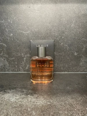 La Rive Heroic Man Eau de Toilette - Fräsch parfym för killar från La Rive, Heroic Man. Flaskan är rektangulär i genomskinligt glas med en silvrig metallkork och ljus orange vätska. Modern och stilren design som passar perfekt i badrumshyllan.