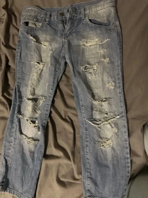  Vintage Capri  jeans med blommor - Säljer ett par blå capri  jeans med kraftiga slitningar och hål framtill. Baksidan har stora broderade blommor i vitt och grönt som ger en unik och kreativ look. Jeansen har klassisk femficksmodell och raka ben. Perfekta för dig som vill sticka ut med din stil. Storlek W27