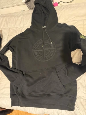 Svart hoodie från Stone Island - Snygg svart hoodie från Stone Island med stor broderad logga på bröstet och klassisk patch på ärmen. Hoodien har dragsko i huvan och en stor magficka. Tillverkad i mjukt bomullsmaterial som känns riktigt skönt mot huden.
