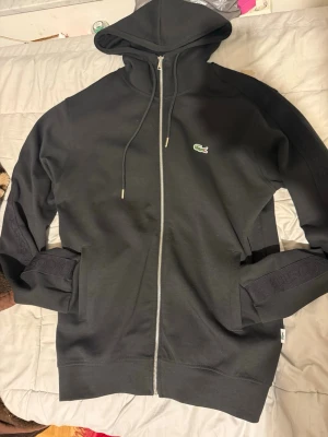 Svart zip hoodie från Lacoste - Snygg svart hoodie från Lacoste med dragkedja framtill och klassisk krokodil-logga på bröstet. Tröjan har huva med dragsko och två fickor på sidorna. Tillverkad i mjukt bomullsmaterial som känns skönt mot huden. Perfekt för en chill och stilren look.