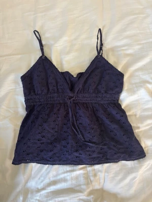 Mörkblå broderad linnetopp - Söt mörkblå linnetopp med broderade detaljer och justerbara smala axelband. Toppen har v-ringning, knyt i midjan och ett luftigt, lätt material som passar perfekt till varma dagar. Broderade hålmönster ger en extra touch.