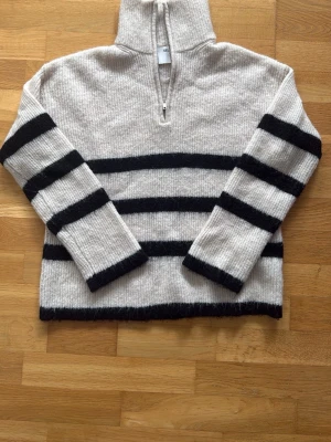 Randig stickad tröja från Selected Femme - Beige stickad tröja från Selected Femme med svarta breda ränder och half zip-dragkedja i halsen. Tröjan har lång ärm och en avslappnad passform, perfekt för kyliga dagar. Ribbad struktur och mjukt material.