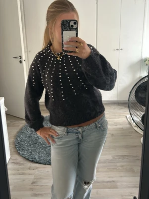 Mörkgrå stickad tröja med pärlor - Säljer en mörkgrå stickad tröja med rund hals och dekorativa vita pärlor som pryder framsidan i ett strålande mönster. Tröjan har långa ärmar och ribbade muddar vid ärmslut och nederkant. Perfekt för dig som vill ha något både mysigt och trendigt.💕