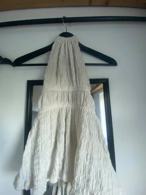 Vit ärmlös halterneck topp - Säljer en vit, ärmlös halterneck topp med smockad struktur och lätt utställd nederdel. Toppen har en luftig och somrig känsla med snygg textur och passar perfekt till både jeans och kjol. Perfekt för dig som gillar en enkel men trendig stil.