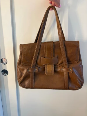 Vintage läderväska -tidlös design  - Snygg brun handväska i skinnimitation med dubbla axelband och dekorativt spänne framtill. Väskan har en klassisk rektangulär form och gott om plats för dina grejer. Perfekt för dig som gillar vintagevibbar och stilren design. Pris kan diskuteras vid snabb affär. 🫶