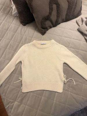 Krämvit stickad tröja med knyt - Säljer en krämvit stickad tröja med rund hals och långa ärmar. Tröjan har ribbade muddar och snygga knytband i sidorna som ger en extra detalj. Perfekt att matcha med jeans eller kjol för en clean look.