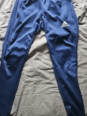 Blå träningsbyxor från Adidas - Säljer ett par blå träningsbyxor från Adidas med smal passform och dragkedjor vid fickorna. Byxorna har Adidas-loggan på vänster lår och är tillverkade i ett mjukt, stretchigt material som passar perfekt till träning eller chill.