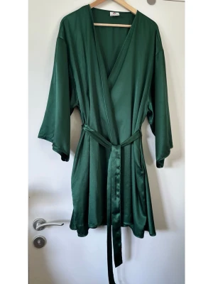 Mörkgrön kimono i satin - storlek L / XL - Elegant stilren kimono / morgonrock / omlottklänning från Abecita i storlek L/XL i en vacker mörkgrön / smaragdgrön nyans. Tidlös modell i omlottdesign med ett matchande knytband i midjan som framhäver figuren. De vida ärmarna ger en bekväm passform och en lyxig känsla. Diskreta fickor i sidorna. Perfekt för avkoppling hemma eller som ett stilfullt plagg över badkläder. Kan även användas som lager över T-shirt och jeans eller till och med som omlottklänning. Lent halvglansigt satin-tyg. 