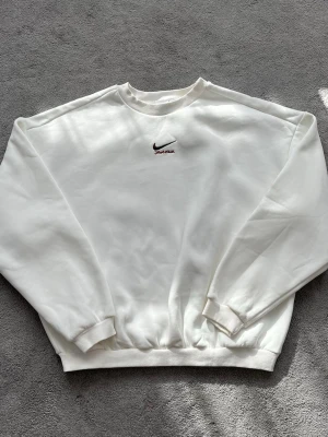 Vit sweatshirt från Nike - Klassisk vit sweatshirt från Nike med broderad svart Swoosh och texten 'Just Do It.' i rött på bröstet. Tröjan har rund halsringning, ribbade muddar vid ärmslut och nederkant samt en avslappnad passform. Perfekt för en clean och sportig look.