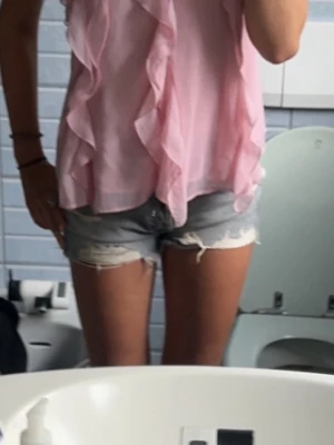 Ljusrosa volangig blus - Säljer en söt ljusrosa blus med volangdetaljer framtill. Blusen är ärmlös och har en luftig, flowy passform i ett tunt, transparent tyg som känns riktigt somrigt. Perfekt att styla med jeansshorts eller kjol för en chill look.
