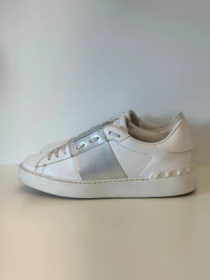 Vita sneakers med silverdetaljer - Snygga vita sneakers med metallic silverdetalj på sidan och nitar längs hälen. Skorna har klassisk låg profil, vita snören och är tillverkade i slätt läder. Perfekta för dig som vill ha en clean men ändå unik look.