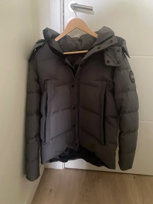 Grå dunjacka från Canada Goose - Säljer en äkta grå dunjacka från Canada Goose med huva och stora framfickor. Jackan har en patch med logga på ärmen och stängs med både dragkedja och knappar. Perfekt för kalla vinterdagar tack vare det tjocka dunfodret och den höga kragen. Fick jackan i present och har använts ett fåtal gånger och är i mycket gott skick, päls och påse ingår!