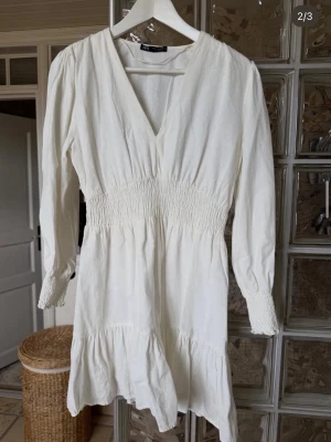 Vit kort klänning från ZARA - Supersöt vit kort klänning från ZARA med v-ringning, långa ärmar och smock i midjan och vid ärmsluten. Klänningen har volang nedtill och är gjord i ett lätt material som känns perfekt för varmare dagar. Storlek M men passar S