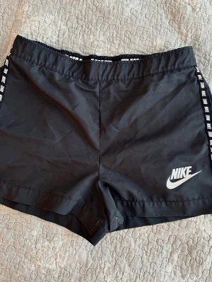 Svarta träningsshorts från Nike М  - Säljer ett par svarta träningsshorts från Nike med elastisk midja och vita loggor längs sidorna. Shortsen har en sportig look och är tillverkade i ett lätt syntetmaterial som passar perfekt för aktiva dagar.