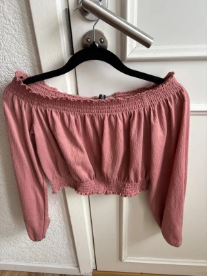 Rosa offshoulder croppad topp - Söt rosa offshoulder topp med lång ärm och croppad passform. Toppen har smockdetaljer vid halsringning och midja samt volangkant upptill och nedtill. Perfekt för dig som gillar romantisk och trendig stil.