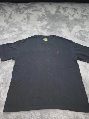 Svart t-shirt från Polo Ralph Lauren - Klassisk svart t-shirt från Polo Ralph Lauren med rund hals och korta ärmar. På bröstet finns den ikoniska röda broderade loggan. Tillverkad i mjuk bomull för en skön känsla och enkel stil.