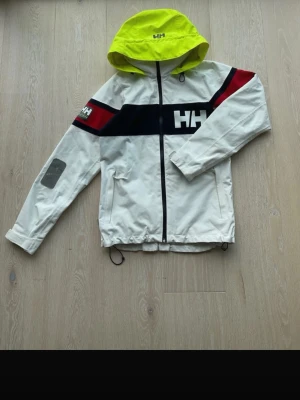 Helly Hansen vit vindjacka med huva - Vit vindjacka från Helly Hansen med neonfärgad huva, svarta och röda detaljer samt HH-logga på bröstet och ärmen. Jackan har dragkedja framtill, två fickor och justerbar nederkant. Perfekt för blåsiga dagar.