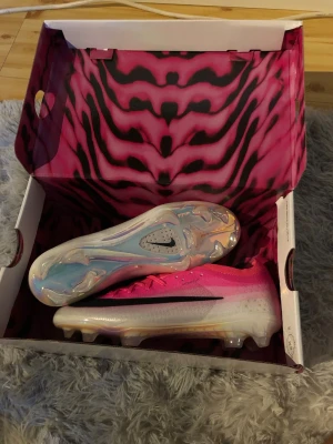 Nike rosa och vita Mercurial fotbollsskor - Säljer ett par Nike Mercurial fotbollsskor i en snygg rosa och vit färggradient med svarta detaljer. Skorna har en låg profil, snörning och en yttersula med dobbar för bra grepp på planen. Materialet är syntetiskt och designen är riktigt modern.